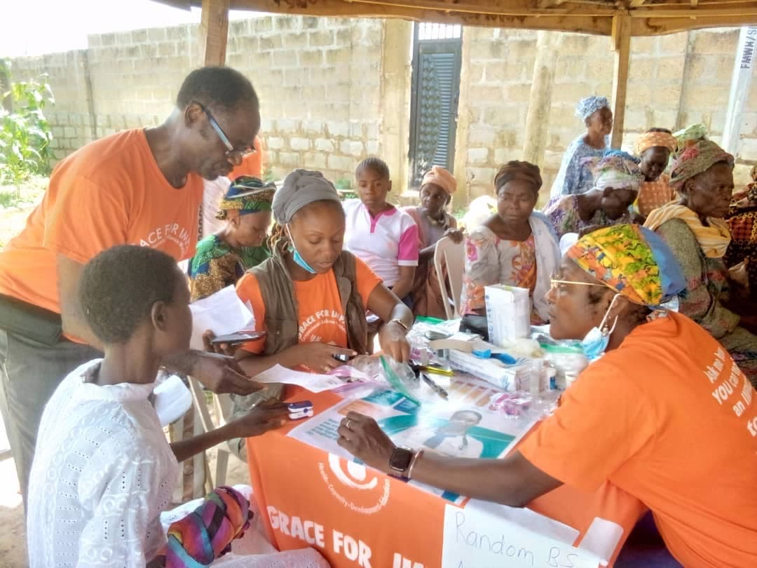 Wide view of an outreach clinic in session, with volunteers in orange shirts attending to a group of community elders seated around tables under an open shelter.