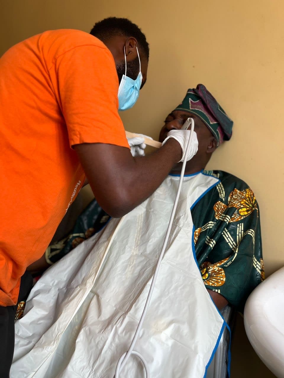 A gloved volunteer attending to a patient in traditional Yoruba attire during a clinic visit.