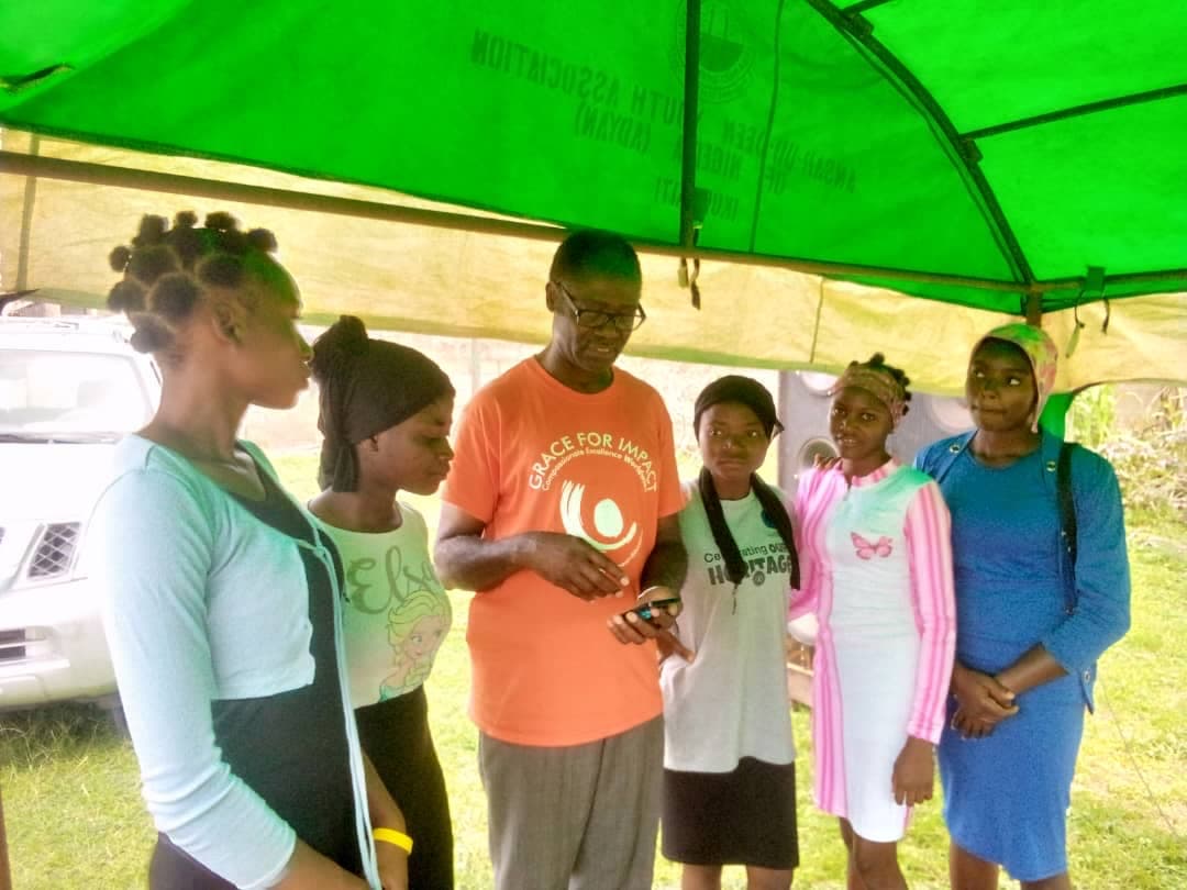 A volunteer in an orange shirt speaking with a group of young women under a green canopy at the outreach.